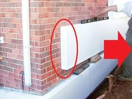 Construction worker installing white siding or paneling on the side of a brick building, with a red arrow pointing to the installation area.