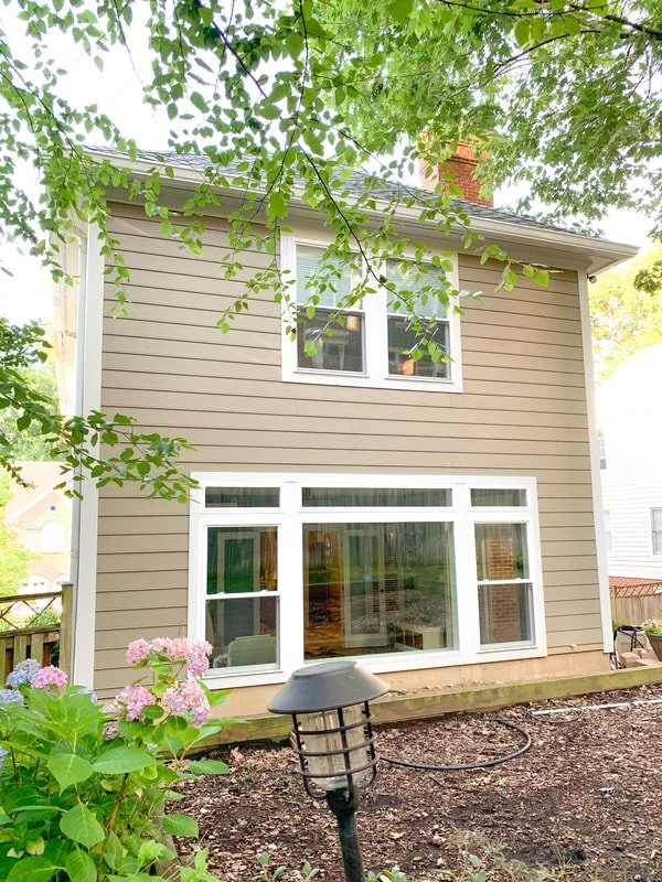 Rear view of a two-story beige house with large white-framed windows, surrounded by green trees and plants, with a garden bed and outdoor light in the yard.