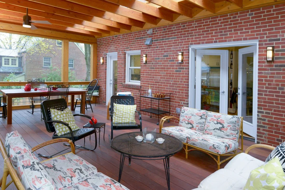 Outdoor porch with brick walls, wooden ceiling, and a screened area, furnished with a sofa, chairs, a round table, and decorative pillows.