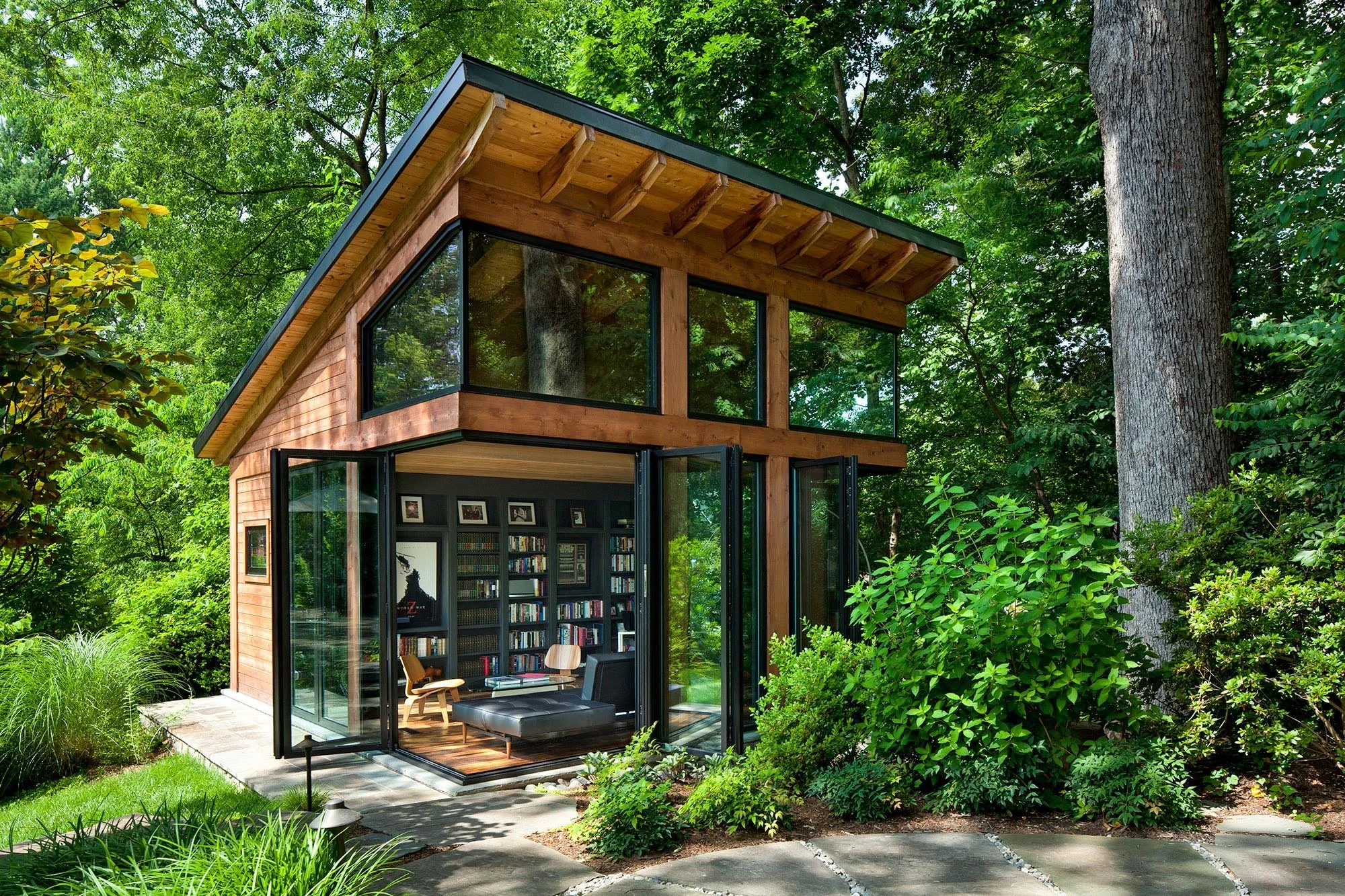 Modern two-story house with wood and glass exterior, surrounded by lush green trees and plants, featuring large windows and an open-concept interior with cozy furniture and bookshelves.
