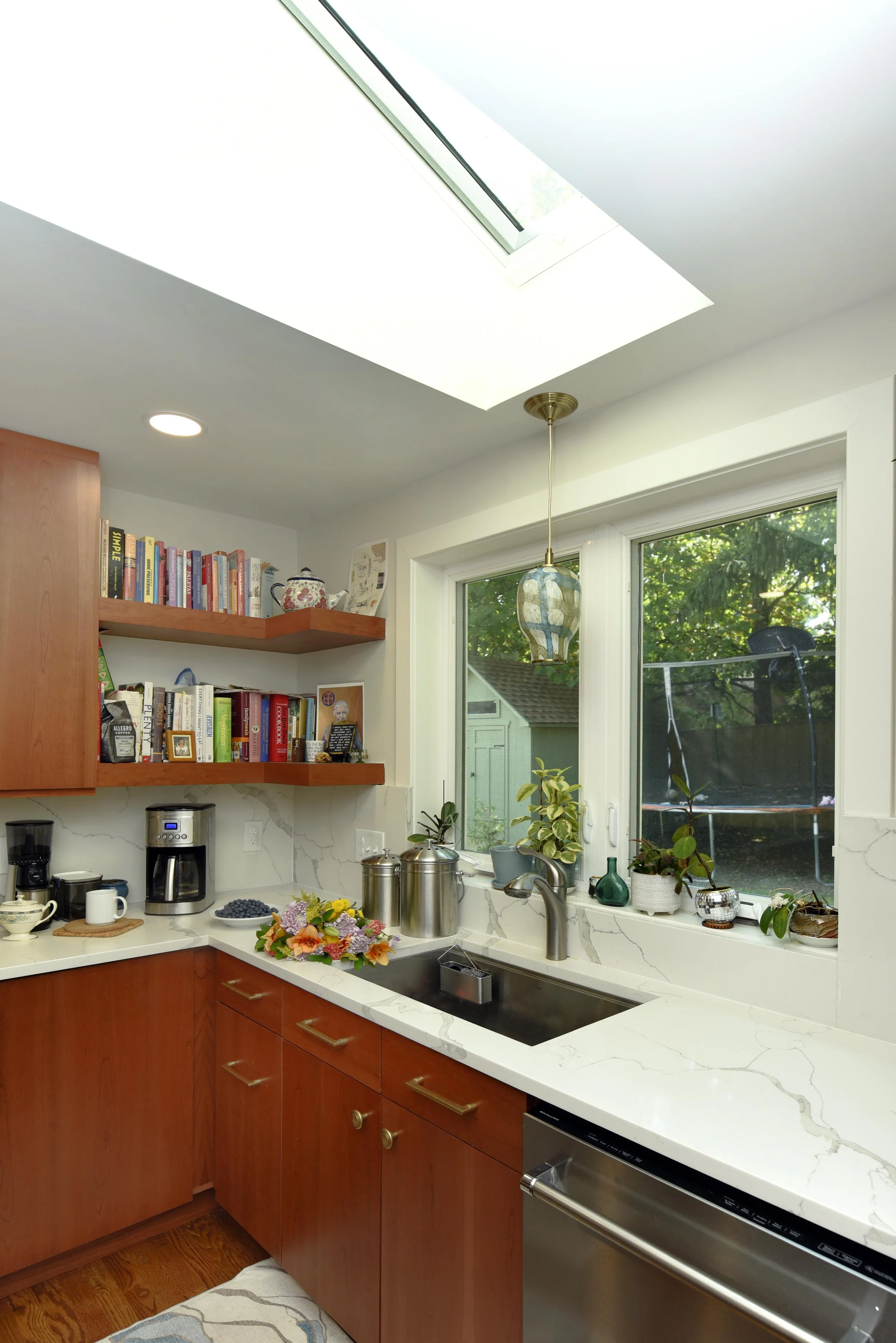 Kitchen with wooden cabinets, a marble countertop, a window with plants, open shelves with books, a coffee maker, and a flower arrangement near the sink.