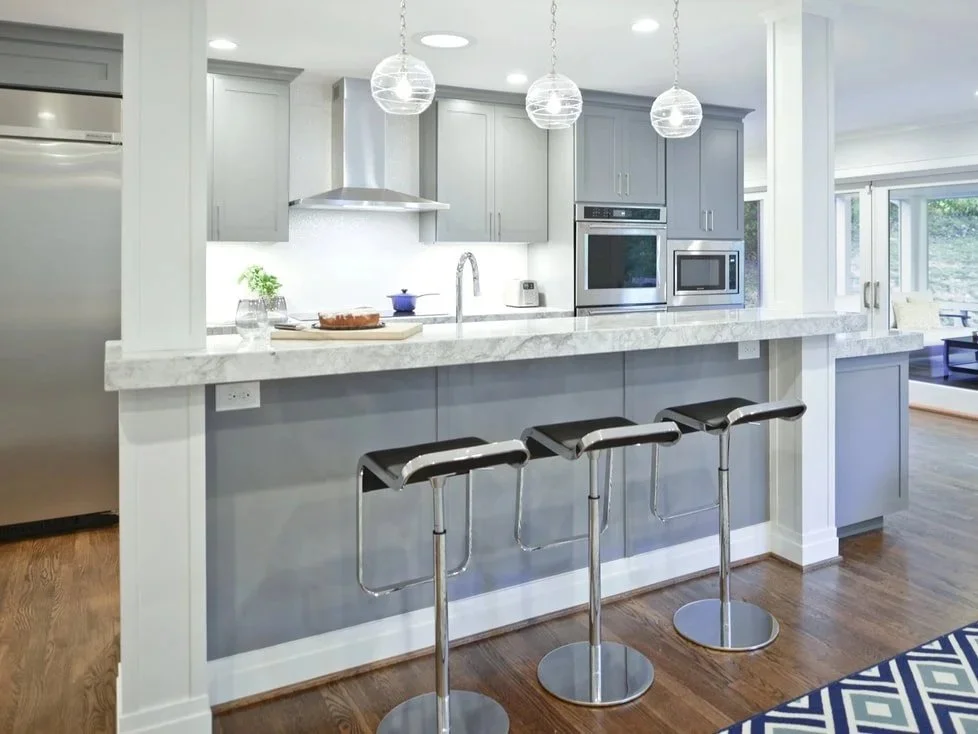 Modern kitchen with light gray cabinets, white marble countertops, stainless steel appliances, and three black barstools at a breakfast bar. Three globe pendant lights hang above the counter.