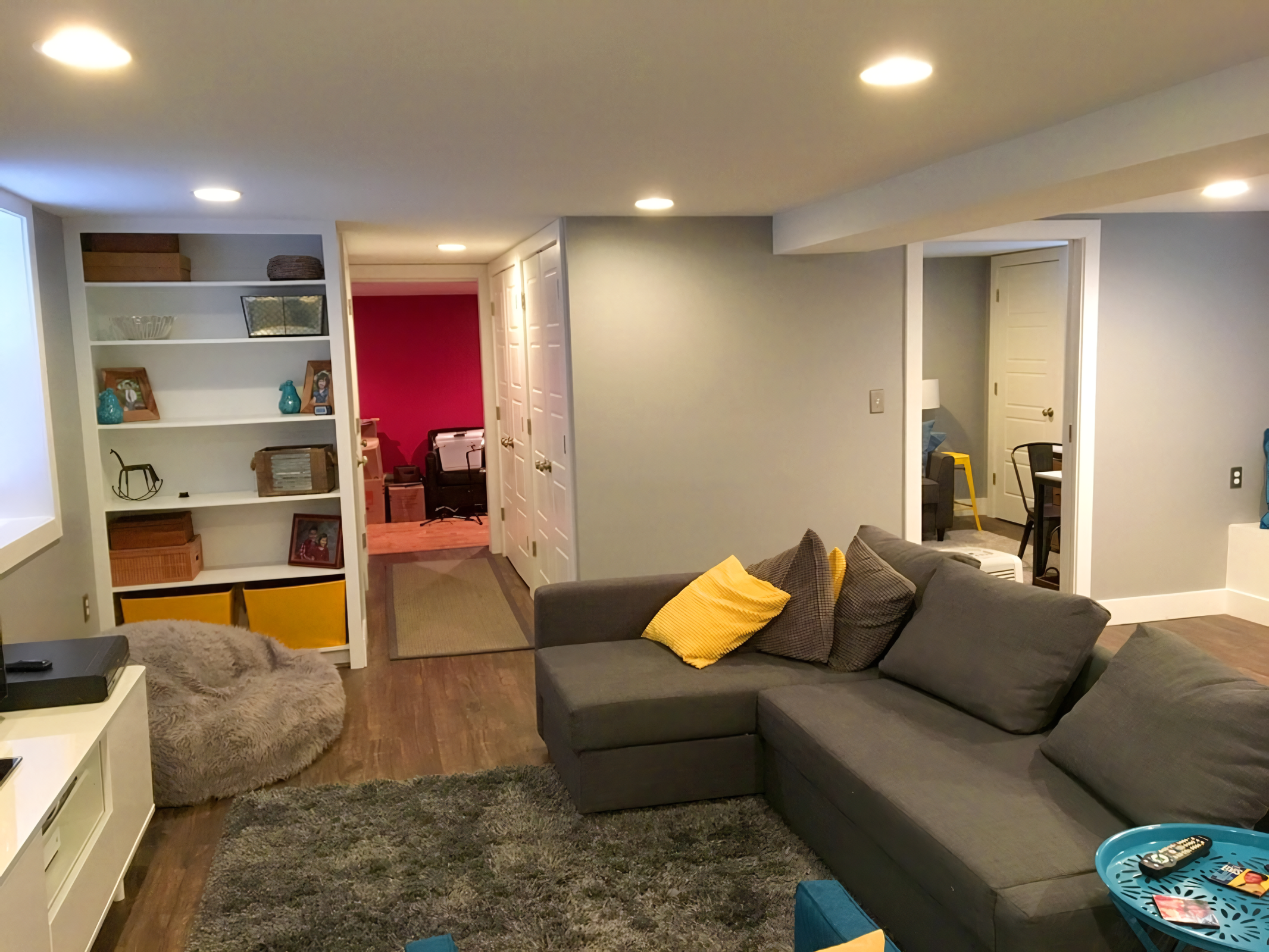Living room with gray sofa, yellow pillows, and a shaggy brown bean bag. A bookshelf with decorative items and photographs. Multiple ceiling lights. Doors leading to different rooms, including a pink-walled office. Wooden flooring and area rugs.