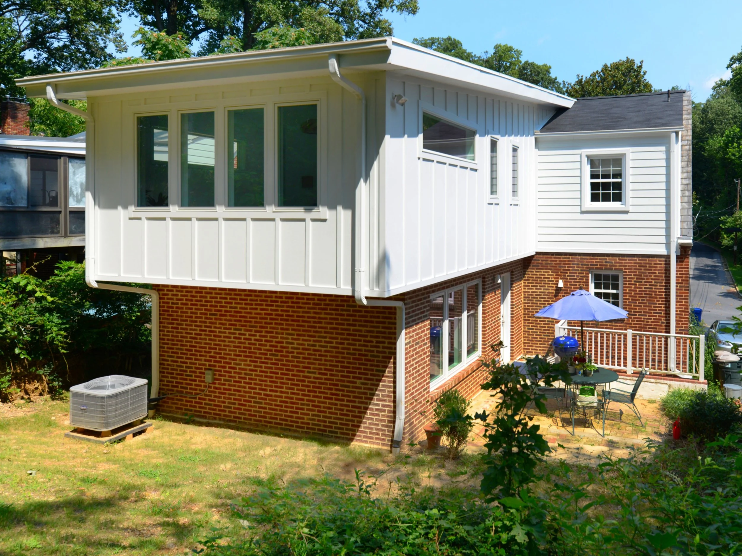 A two-story house with white wooden siding and a brick foundation, surrounded by green trees and a backyard with a patio, grill, and patio furniture.