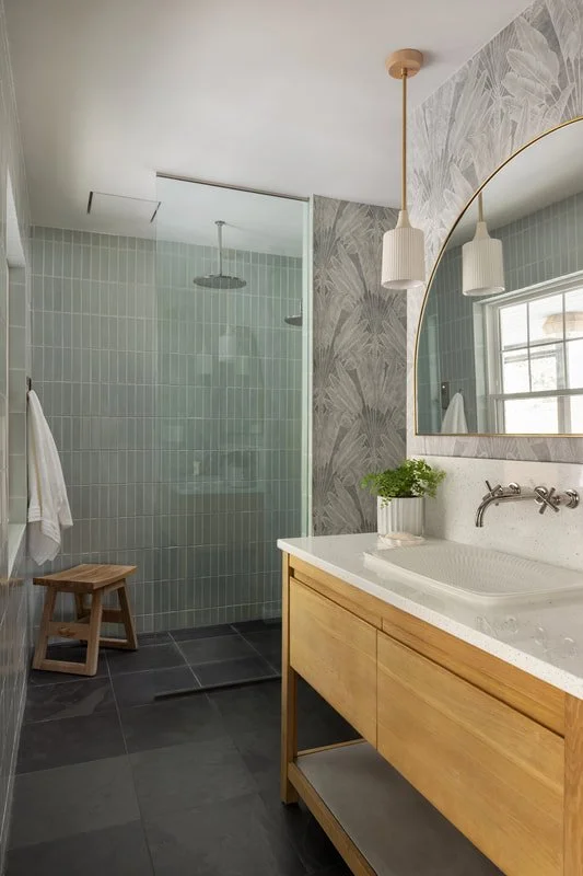 A modern bathroom with a glass shower enclosure, wooden vanity with a white sink, large mirror, and wall-mounted faucet. There's a potted plant on the vanity, a wooden stool, and a towel hanging on a hook. The bathroom features light-colored walls wi