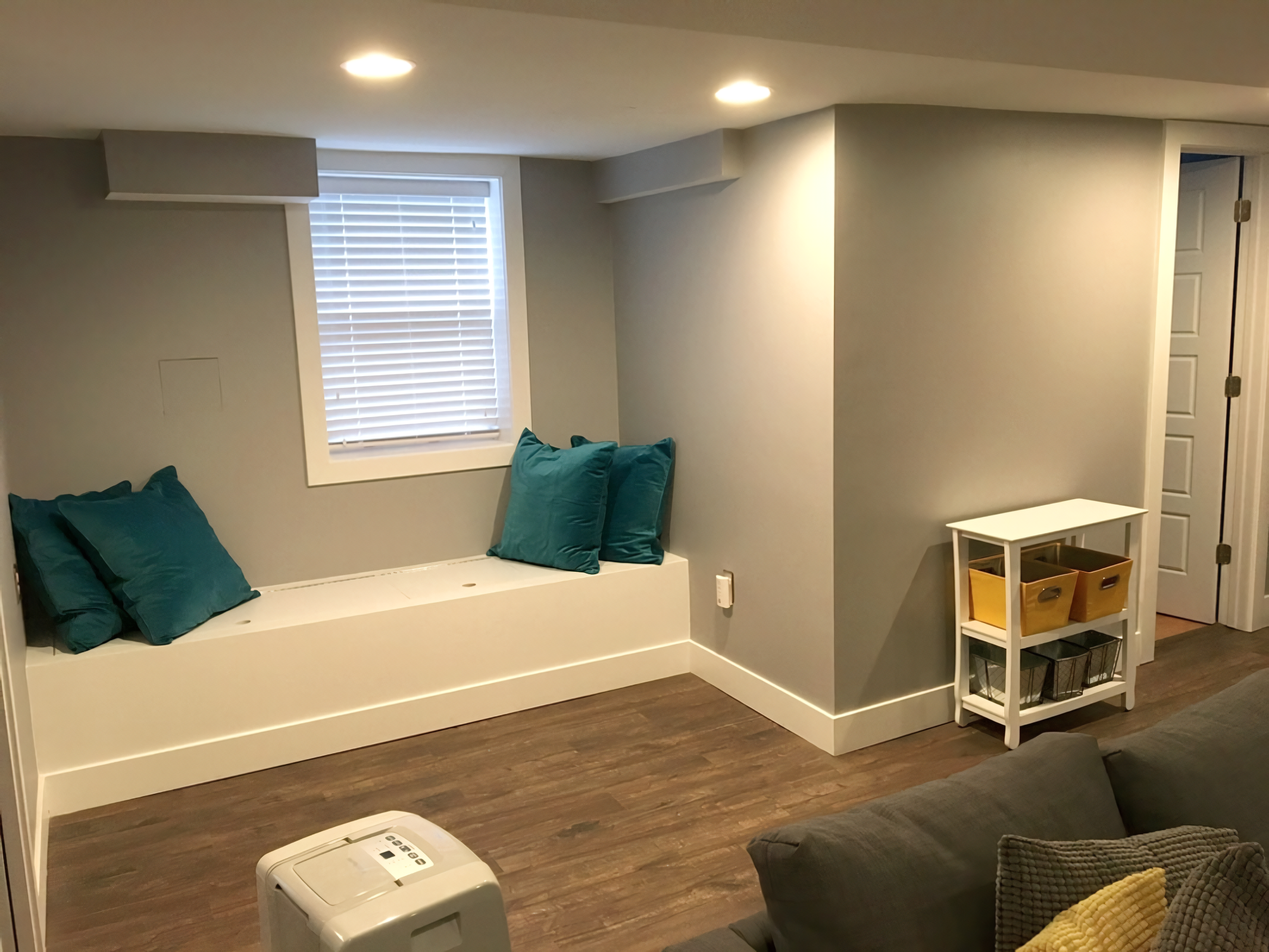 Living room with a window seat with teal pillows, a window with blinds, a small white storage shelf with yellow and black baskets, a gray sofa with cushions, and a portable air conditioner in a room with hardwood floors.