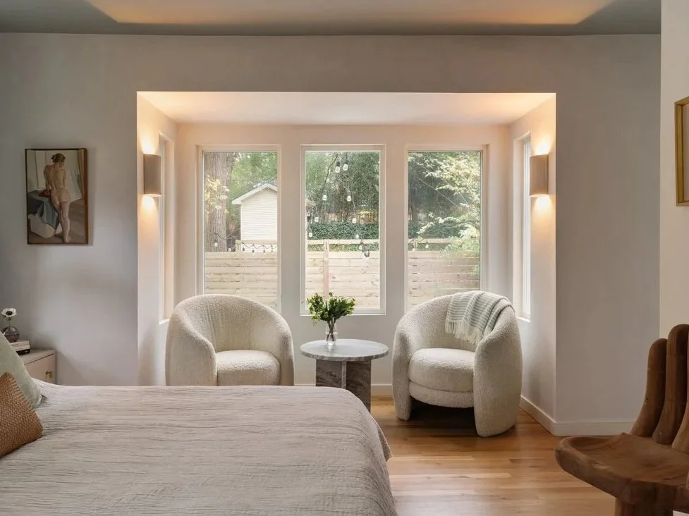 A cozy bedroom corner with two white upholstered armchairs and a small round marble table with a plant, by a large window with a wooden fence and garden outside. The room has wall-mounted lights and minimal decor.