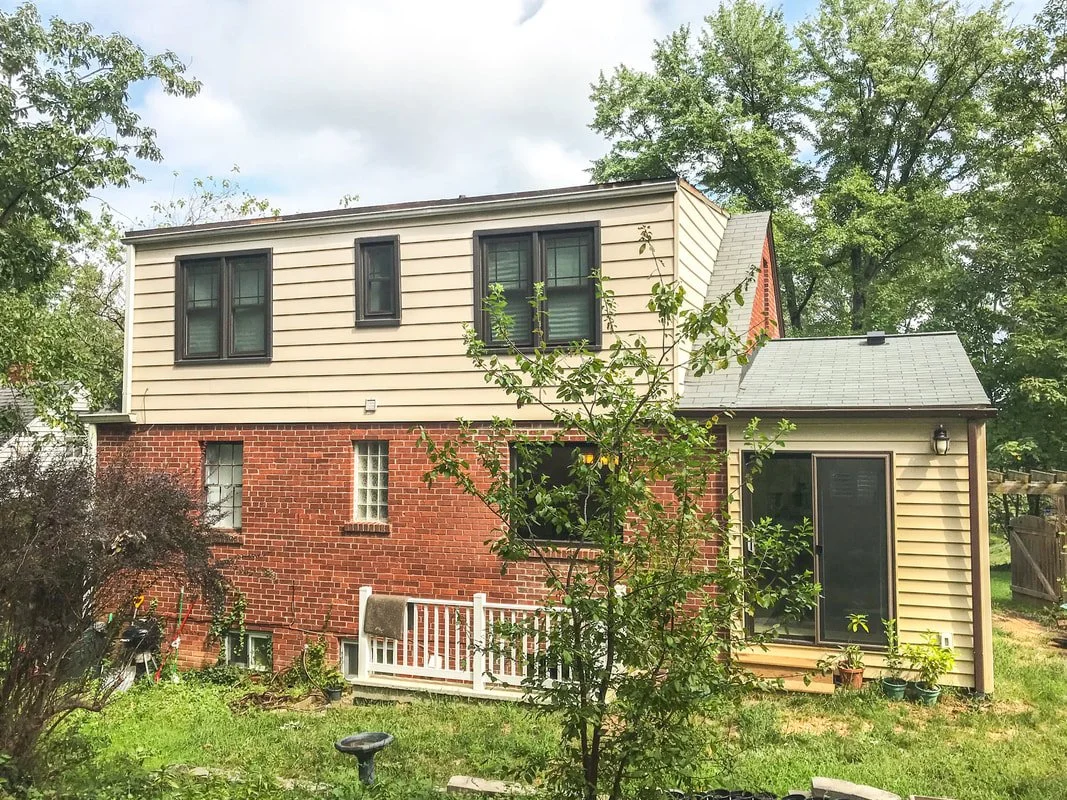 A two-story house with a brick lower level and beige siding upper level, with multiple black-framed windows. The yard has trees, bushes, and a small white fence.