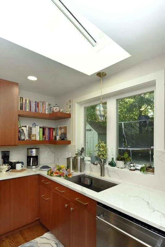 Bright kitchen with wooden cabinets, white marble countertops, open shelves with cookbooks, a coffee maker, and plants near a window.