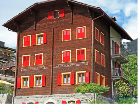 A three-story wooden house with red window shutters and a stone foundation, surrounded by greenery, under a partly cloudy sky.