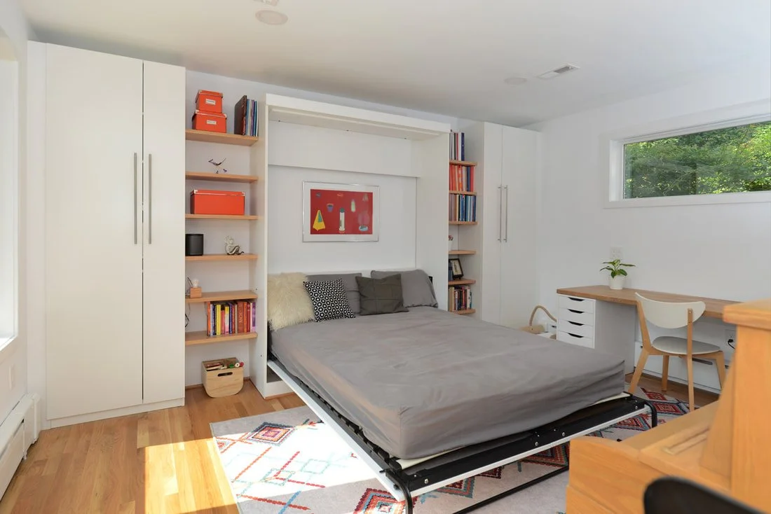 A bedroom with a bed frame and mattress, white walls, a window with green trees outside, built-in white closets, a small desk with a chair and a potted plant, and open shelves with books and storage boxes.