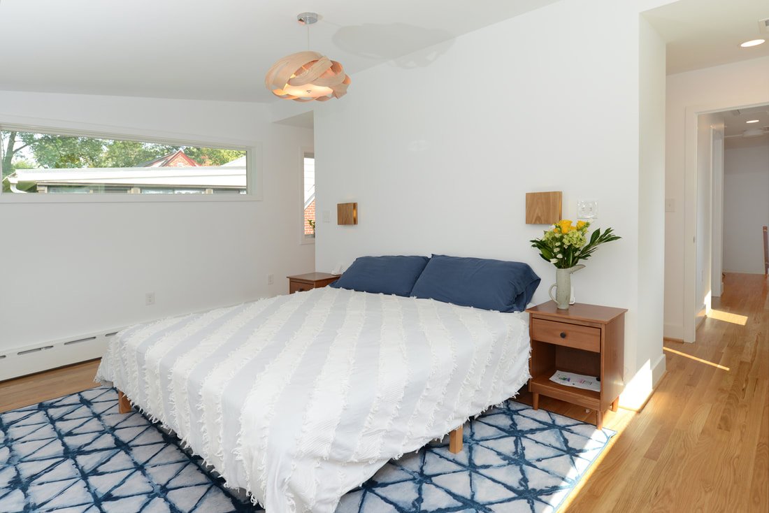 A bedroom with a large bed, white textured bedspread, navy pillows, wooden nightstands, a vase of yellow flowers, a patterned blue and white rug, and a modern ceiling light fixture.