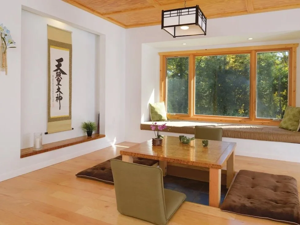 A cozy, sunlit room with wooden floors, a large window seat with green cushions, a low wooden table, two cushioned floor chairs, and Asian-themed wall decor.