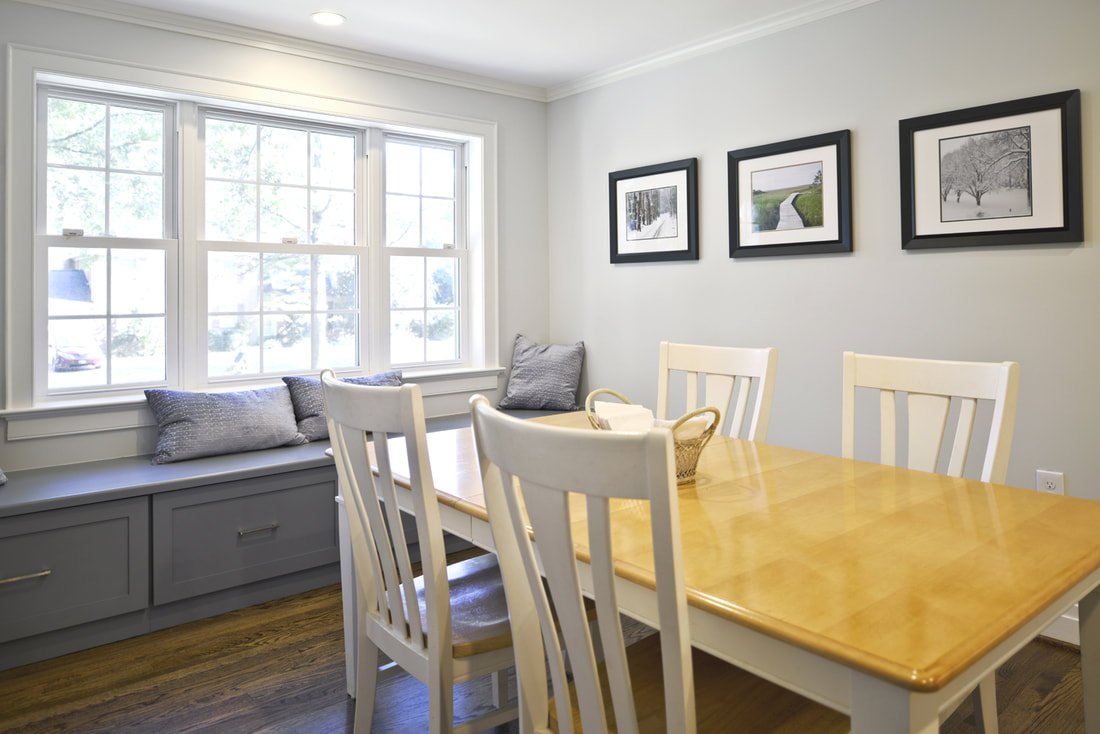 Dining room with a wooden table, white chairs, window seat with pillows, and framed black-and-white landscape artwork on the wall.