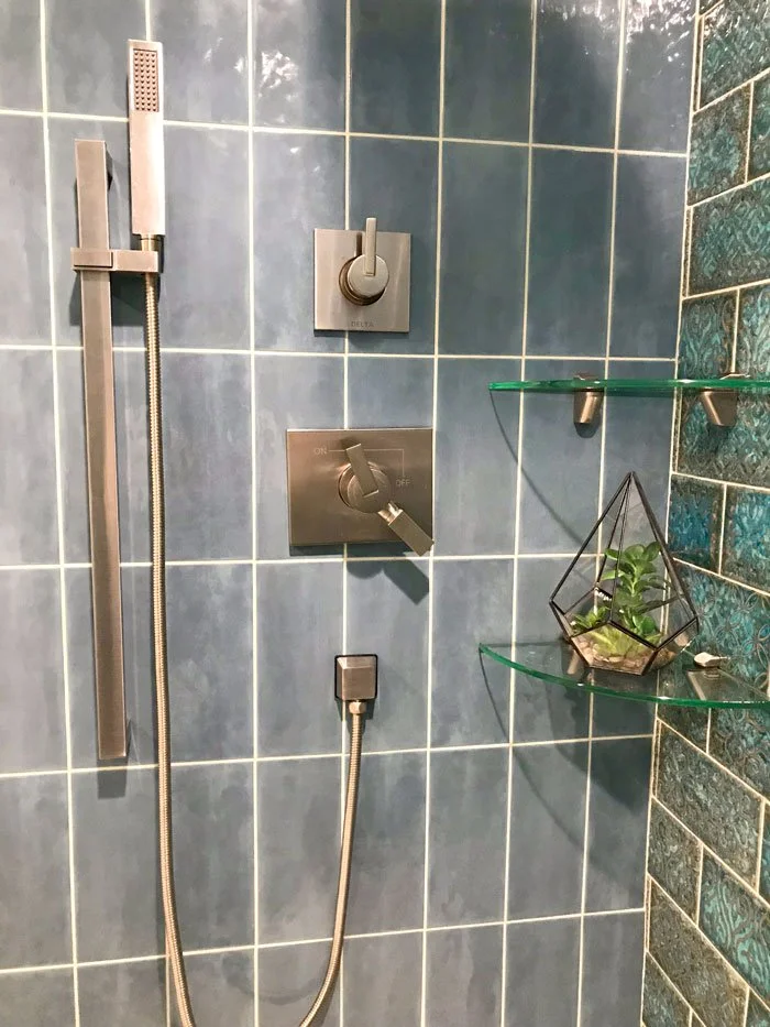 Shower with gray tiles, a handheld showerhead, a control panel with a switch, and a glass shelf with a plant in a geometric holder.