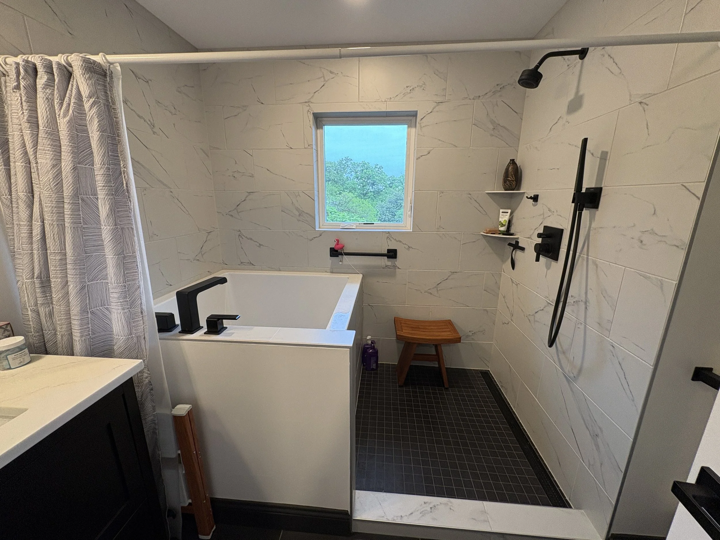 A modern bathroom with white marble walls, a walk-in shower with a black showerhead, a window with a view of greenery, a small shelf with a decorative vase, a pink flamingo towel hook, a wooden stool, and a black and white checkered rug.