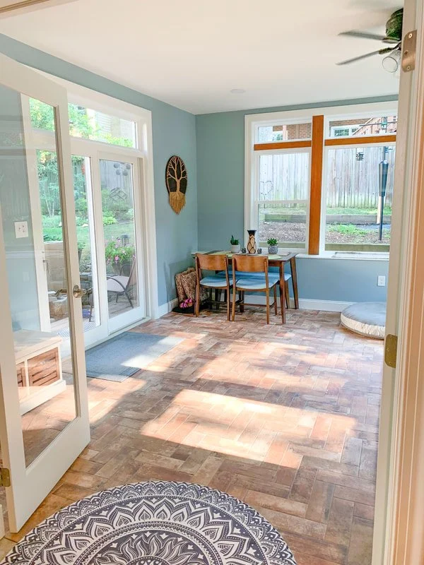 Bright, sunlit dining area with blue walls, wooden floor, glass sliding door opening to patio, and a table with three chairs near a large window. Decor includes wall hanging and small plants.