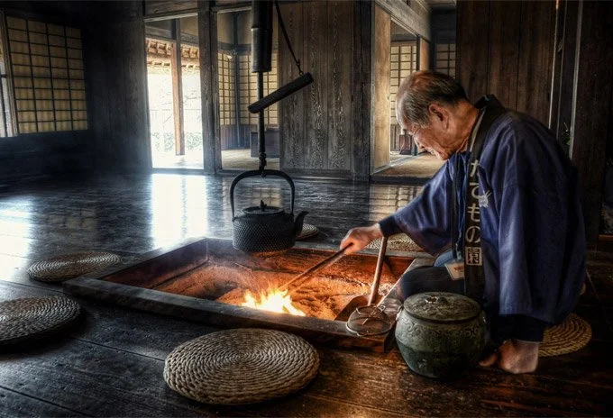 A man in traditional Japanese attire is sitting on the floor, using a tool to stoke a fire inside a large, rectangular iron furnace, with a kettle hanging over it and traditional woven cushions around him in a historical Japanese room.