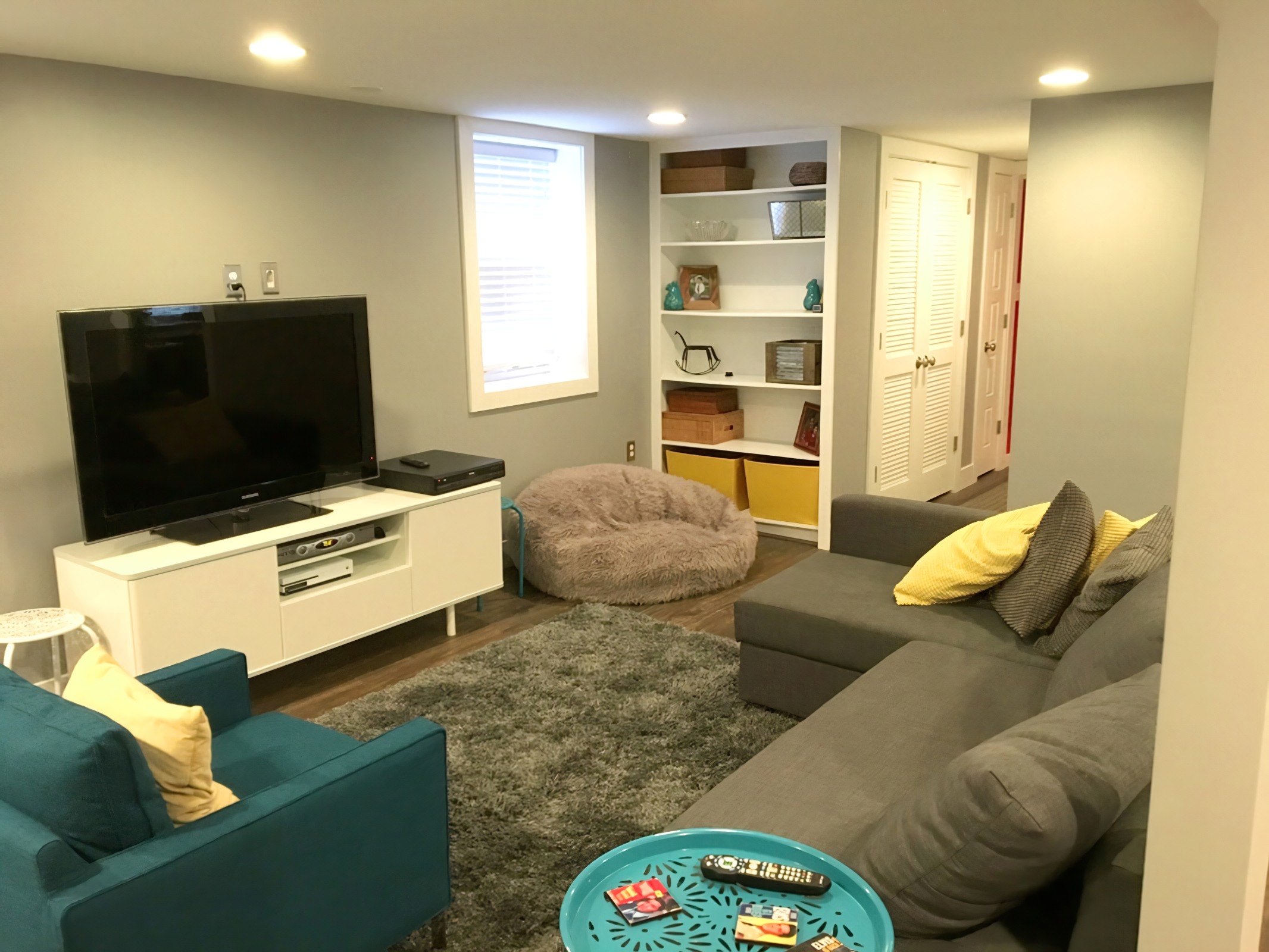 A cozy living room with a gray sectional sofa, a teal armchair, a TV on a white stand, a black TV remote, a fluffy bean bag chair, a patterned area rug, and various decorative items on a white bookshelf. There is a window with blinds and a hallway with closed white doors.