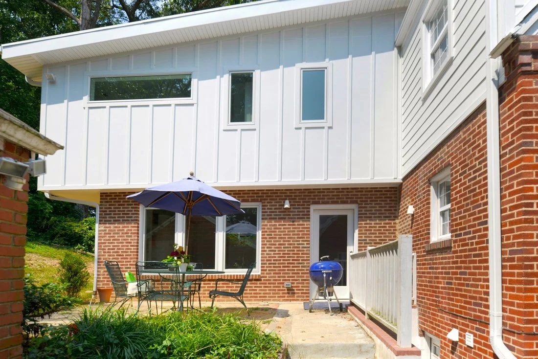 Backyard patio with a round table, chairs, an umbrella, a small grill, and a sliding glass door, with a two-story house featuring brick and white siding.