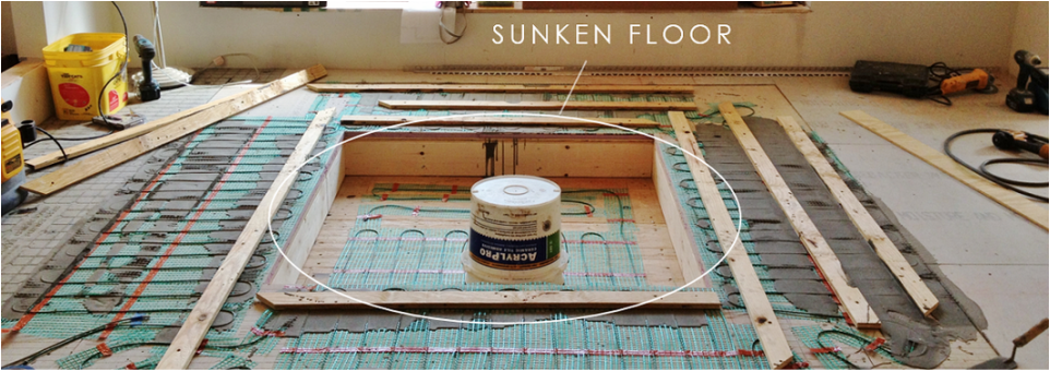 Construction site showing a sunken floor with exposed subfloor and wooden framing, surrounded by construction tools and materials.