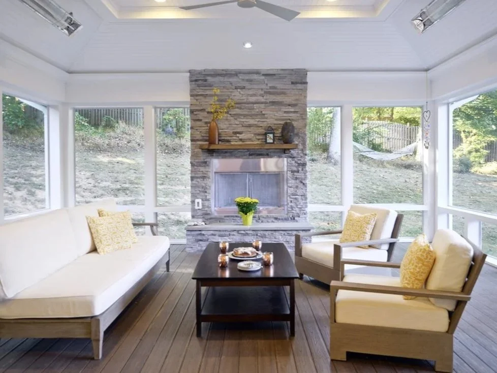Bright sunlit room with large windows, a stone fireplace with a wooden mantel, a vase with yellow flowers, a cream sofa with yellow pillows, and two armchairs with matching pillows, wooden floor, and outdoor greenery visible through windows.