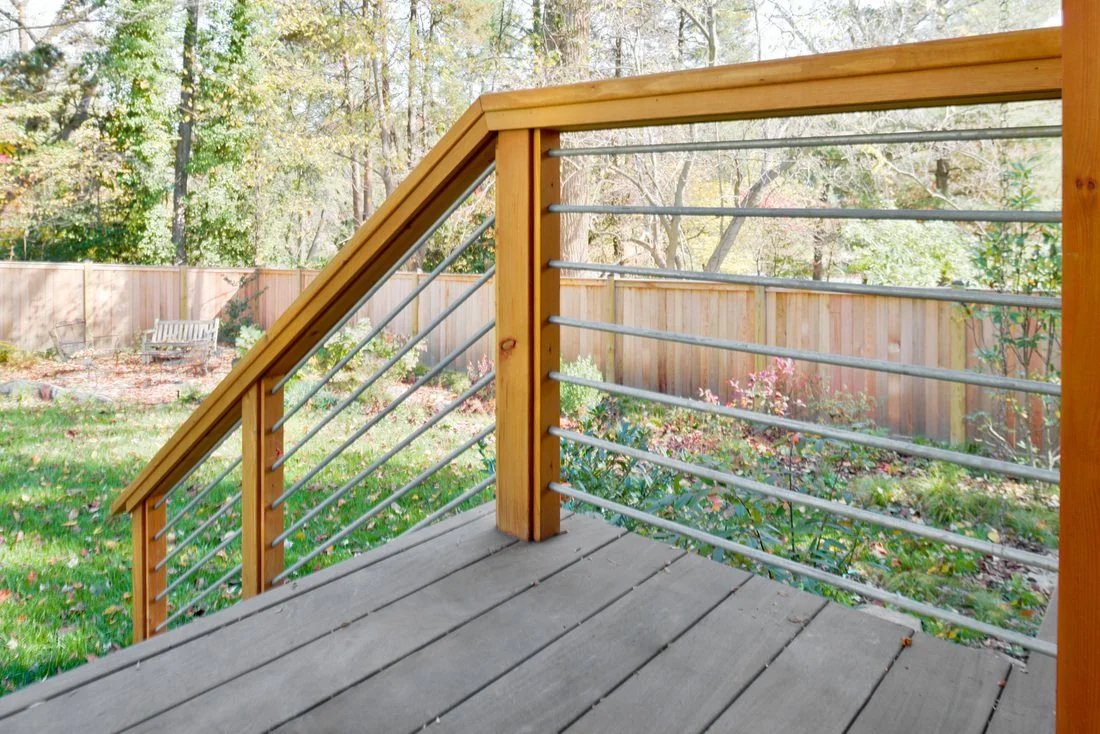 View of a backyard deck with wooden railing and metal cables, overlooking a garden with trees and a wooden fence.