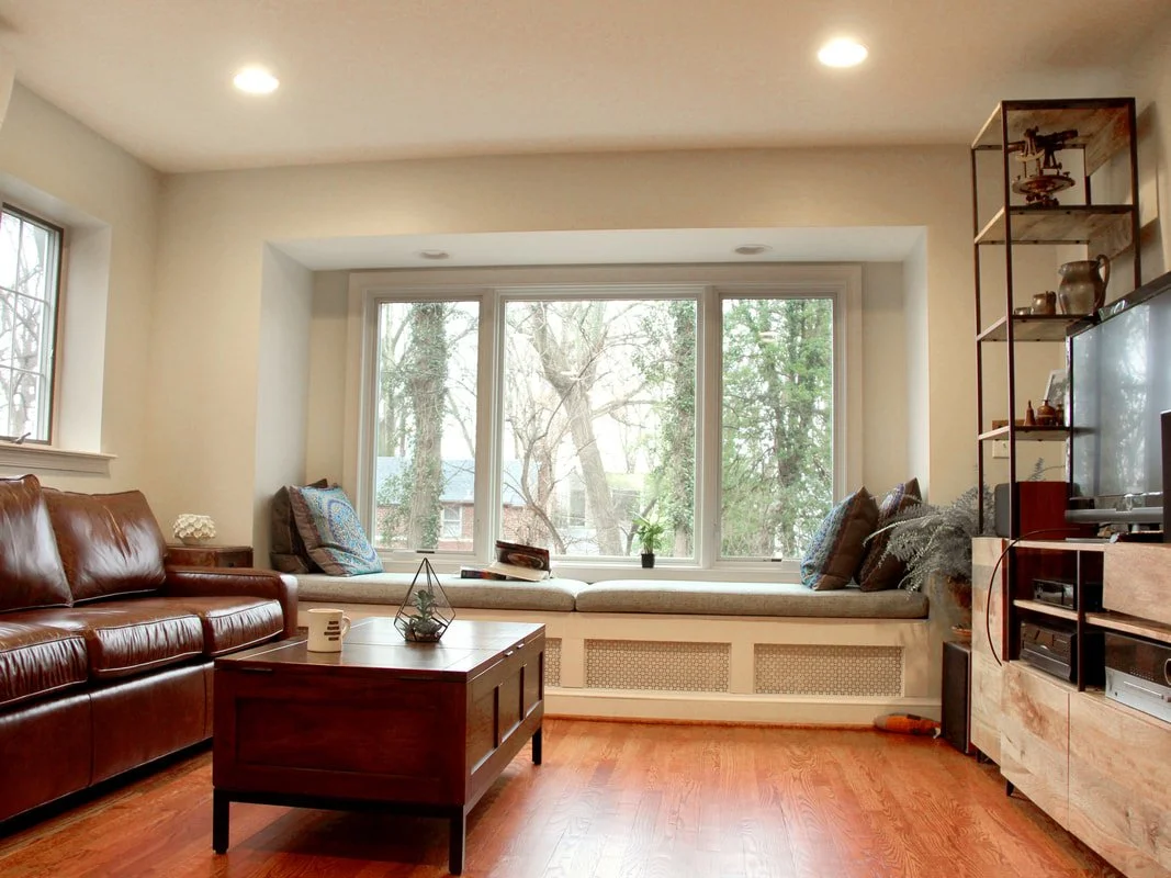 Cozy living room with a large window seat, leather sofa, wooden coffee table, and shelves with decorative items.