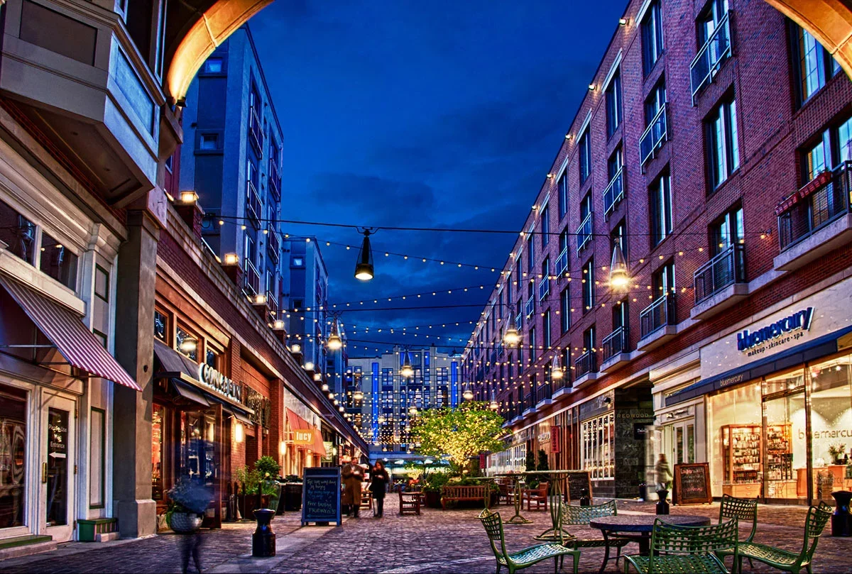 An outdoor shopping and dining area illuminated with string lights at dusk, featuring shops, a tree, and people walking.