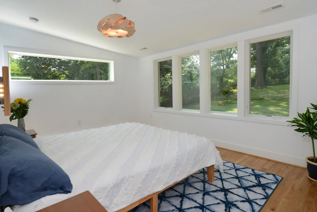 Bedroom with large windows, white walls, ceiling light fixture, bed with white bedding and blue pillows, nightstand with yellow flowers, blue and white patterned rug, and a potted plant near the window.