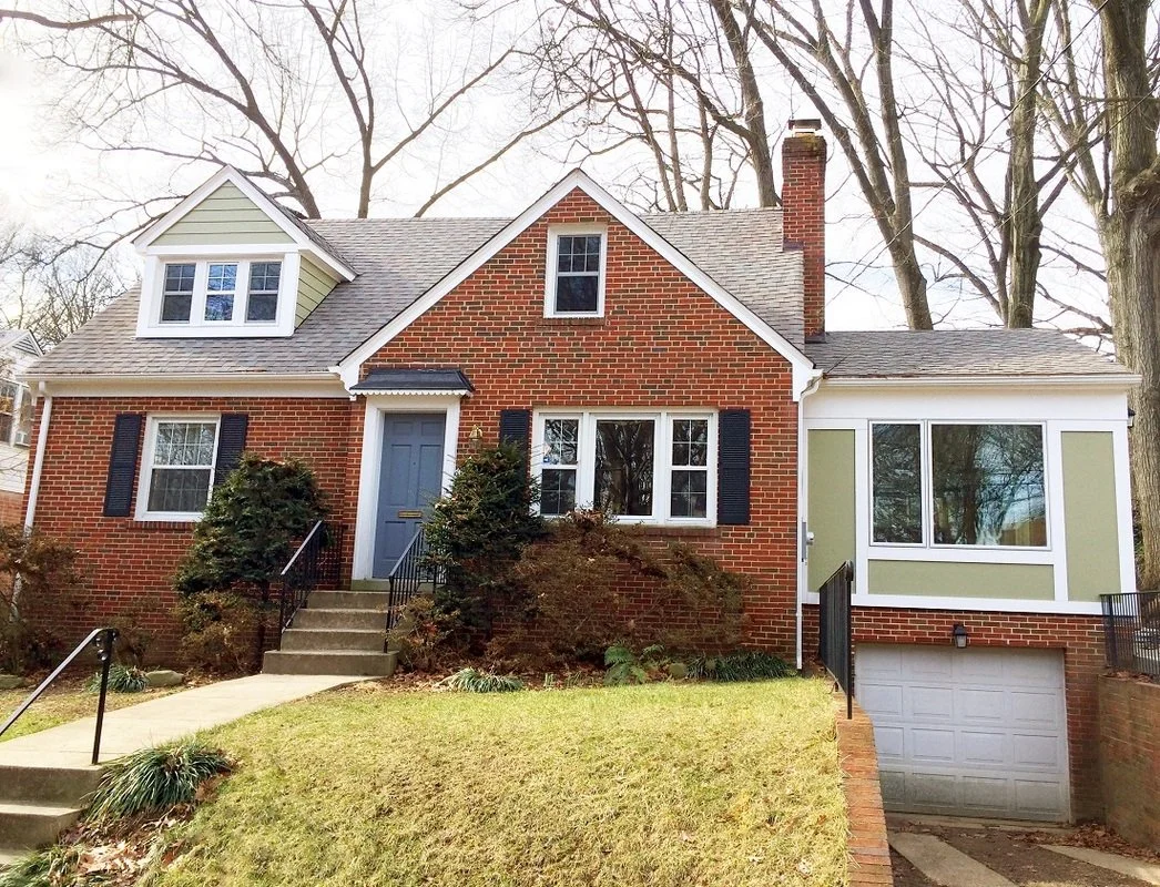 A two-story brick house with a green garage door, white window trims, black shutters, and a small front porch with steps leading up to the front door. There are leafless trees in the background and a sloped lawn in the front.