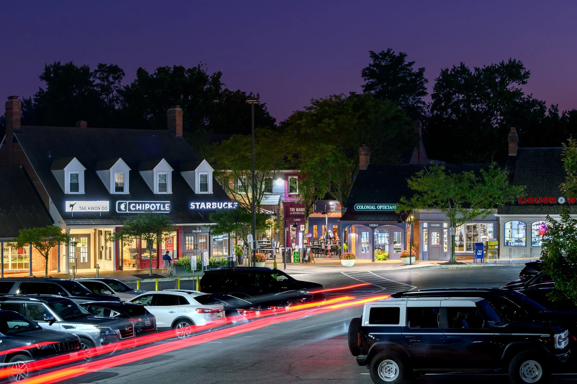 Night view of a lively shopping street with various storefronts including Chipotle, Starbucks, and Colonial Opticians, with parked cars in the foreground and trees lining the street.