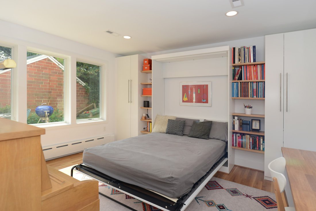 A bedroom with a bed that has an adjustable base, flanked by built-in white bookshelves filled with books, and three large windows showing an outdoor yard with a brick wall and a blue grill.