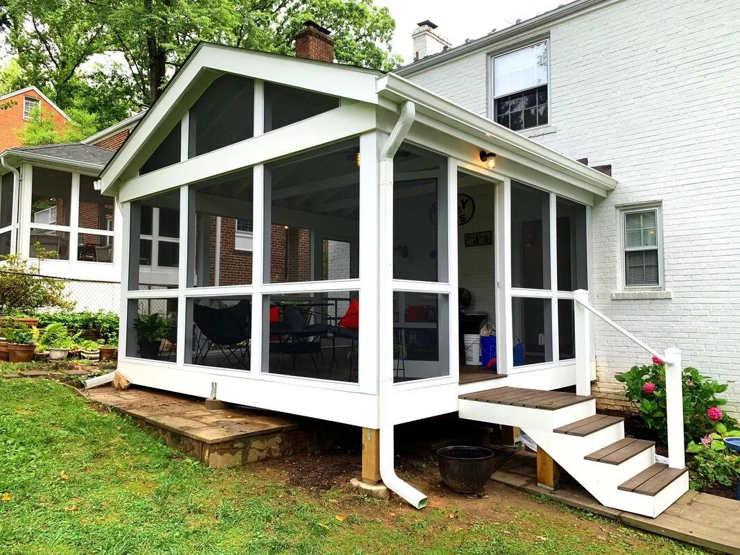 A white screened porch attached to a white brick house with wooden steps leading up to the porch, surrounded by a grassy yard and flowering plants.