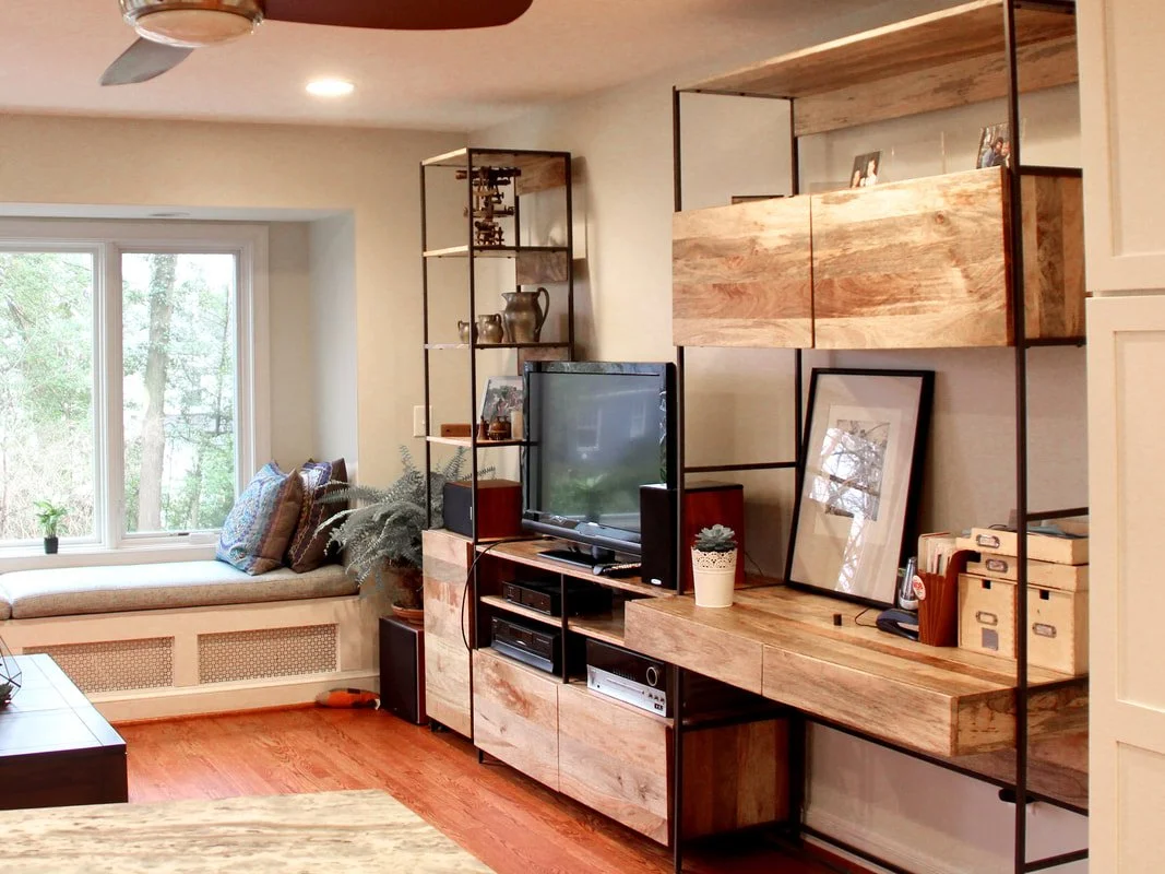 Living room featuring a cozy window seat with pillows, a TV on a modern wooden and metal entertainment unit, and a large window with trees outside.