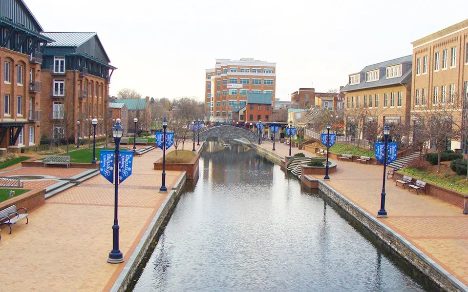 A canal flanked by brick buildings and a pedestrian walk with benches, street lamps, and blue banners, with a small bridge in the distance.