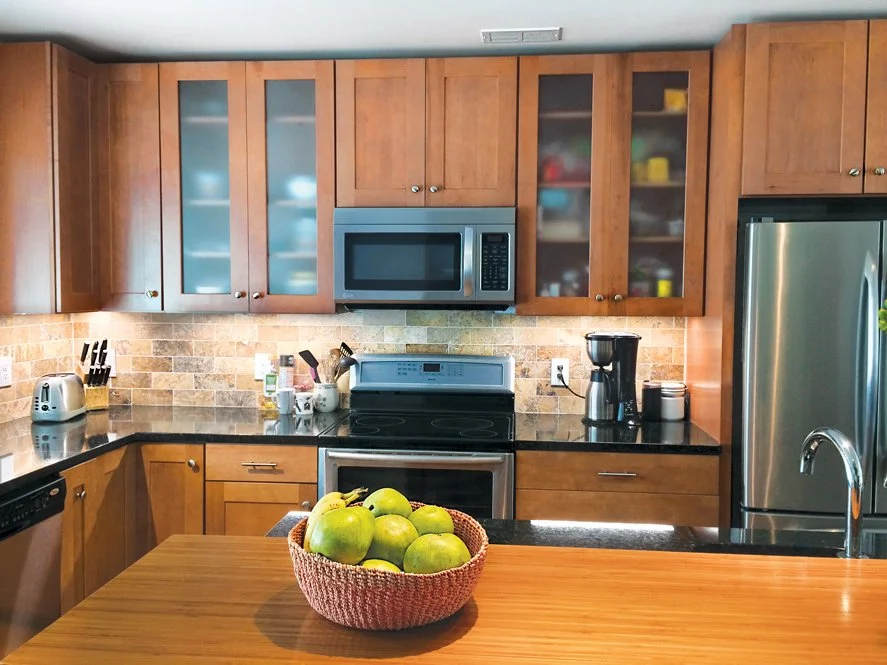 A kitchen with wooden cabinets, a microwave above the stove, a stainless steel refrigerator, and a wooden countertop with a bowl of green apples.