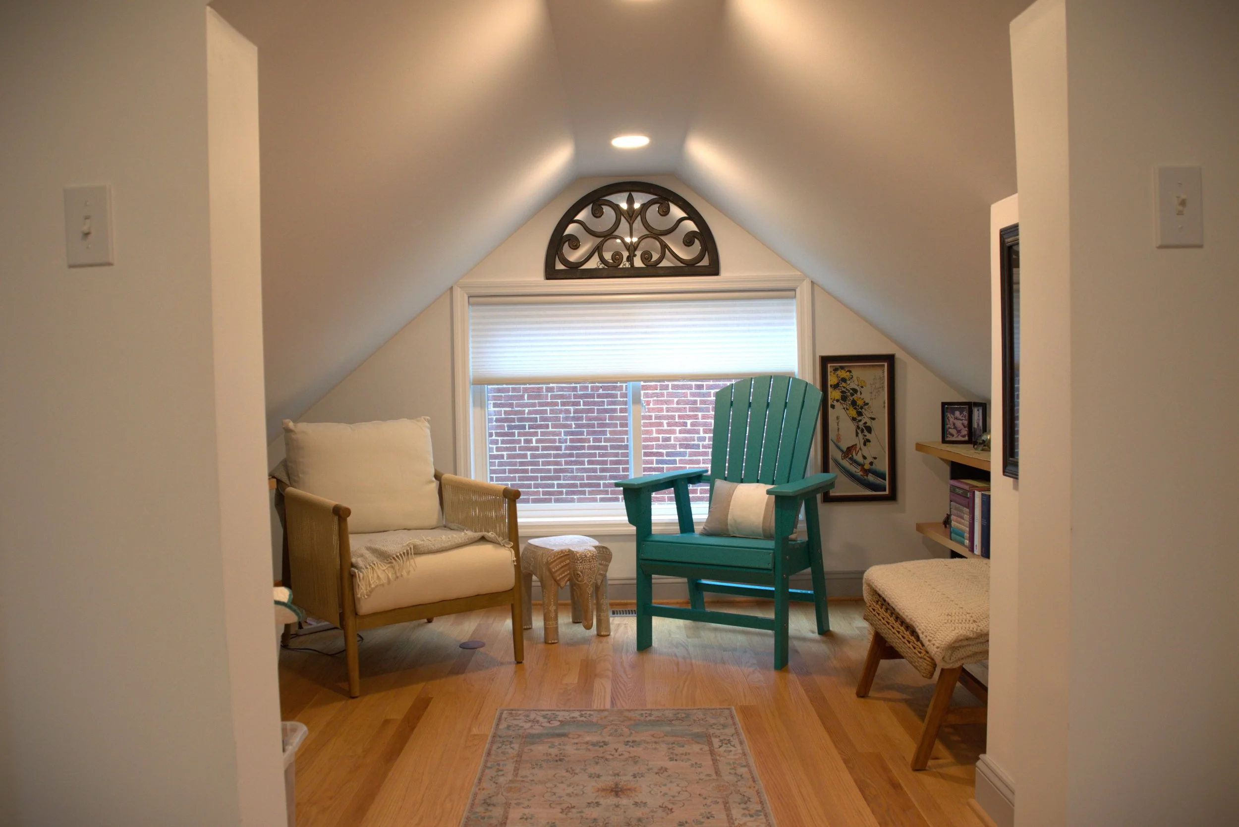A cozy attic room with sloped ceilings featuring a window with a brick wall visible outside. The room is furnished with a cream armchair with a blanket and cushion, a turquoise Adirondack chair, a small patterned stool, a beige woven bench, and frame