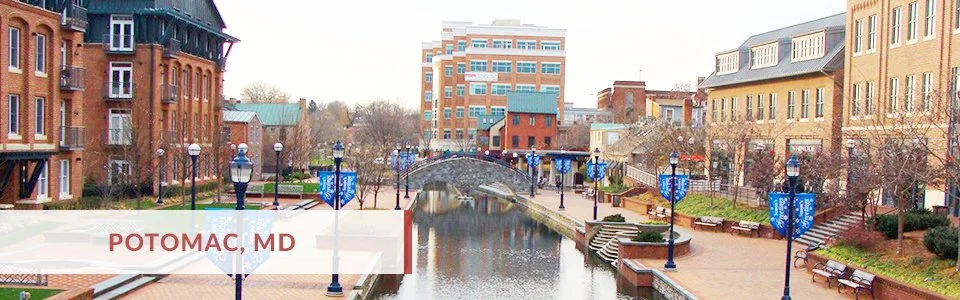 Cityscape view of Potomac, Maryland, featuring a canal with a small arched bridge, surrounding office and residential buildings, paved walkways, benches, and street lamps with blue banners.