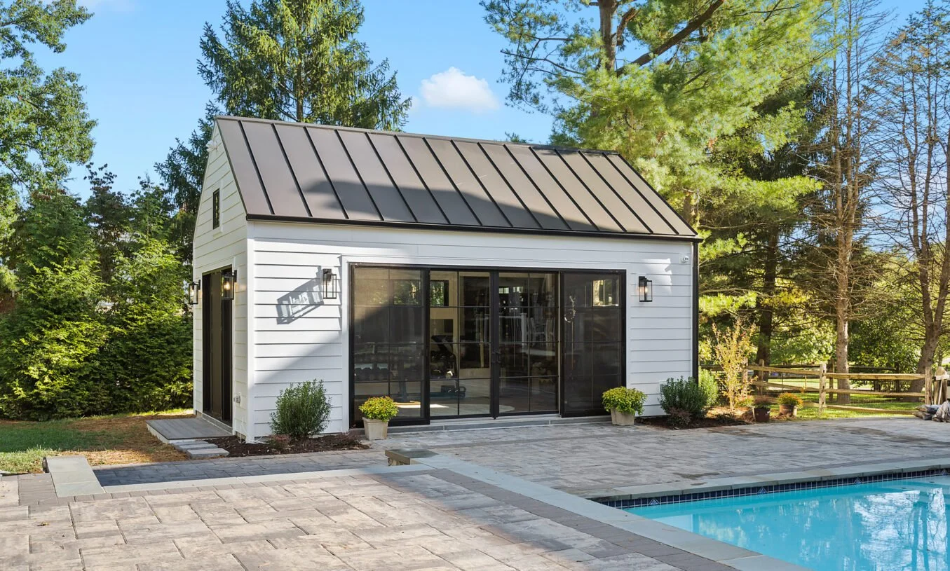 White pool house with black sliding glass doors, surrounded by trees, with a swimming pool in the foreground, and a patio with paving stones.