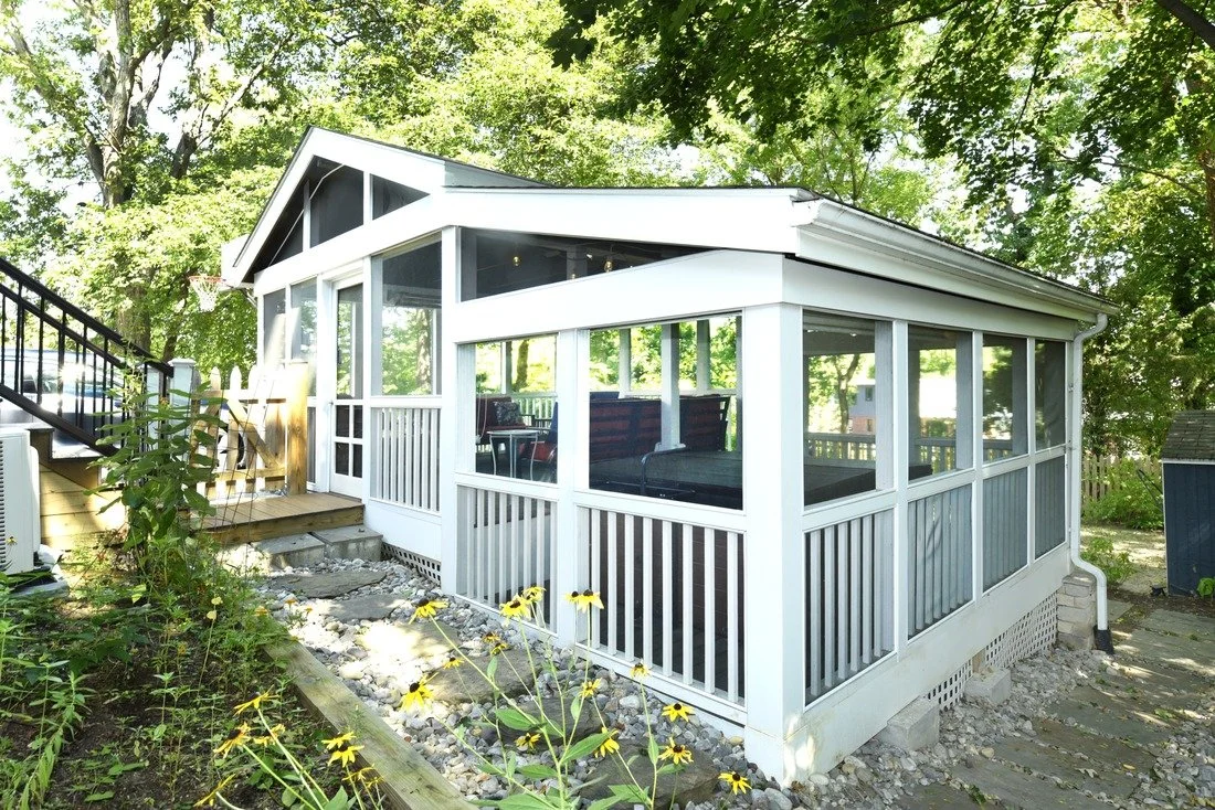 A screened-in porch with white framing attached to a house, surrounded by green trees, with a small flower bed and a stone pathway in front.