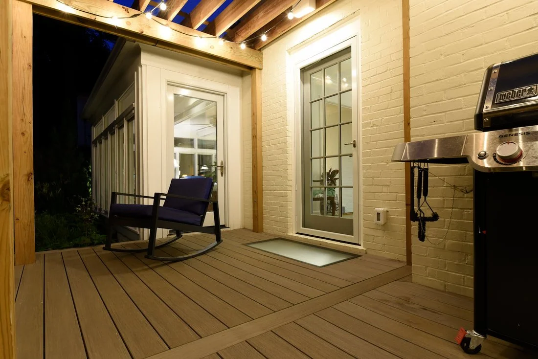 Covered outdoor patio with wooden flooring, a rocking chair, string lights overhead, a side door, a glass-paneled door, a brick wall, and a grill.
