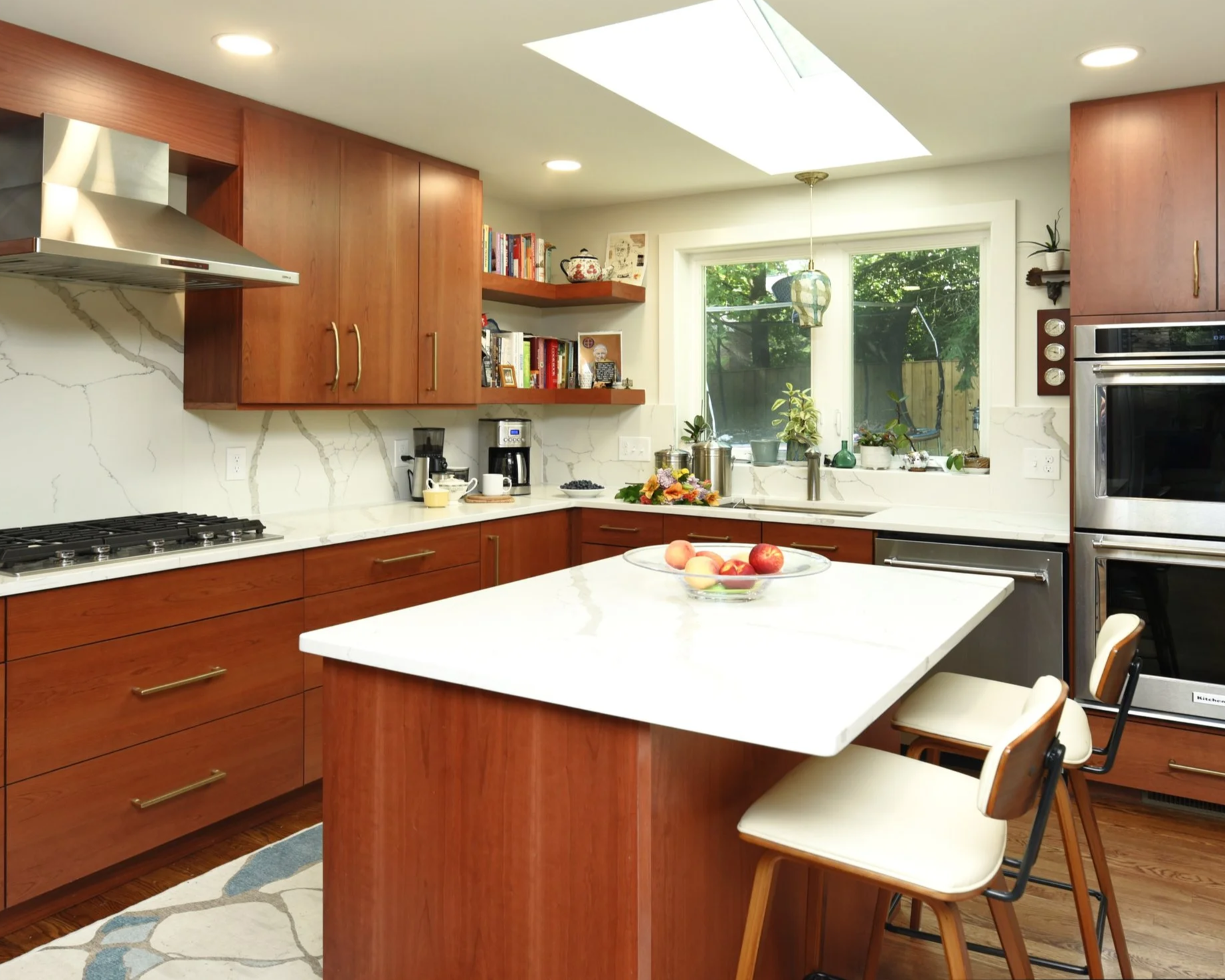 A modern kitchen with wooden cabinets, a white marble backsplash, a central island with a white countertop, and a window showing green outdoor scenery. The kitchen features built-in appliances, open shelves with books, and a bowl of apples on the island.