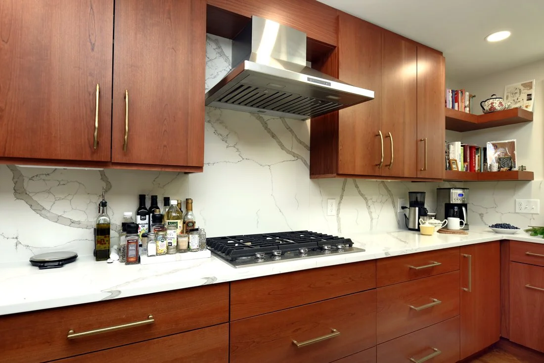 Kitchen with wooden cabinets, a marble backsplash, a gas stove, and various kitchen appliances and bottles on the counter.