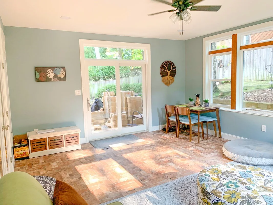 Bright living room with blue walls, hardwood floor, sliding glass door leading to a backyard patio, a small dining table with two chairs, and large windows with wooden crossbars. A ceiling fan with attached lights hangs from the ceiling, and decor in