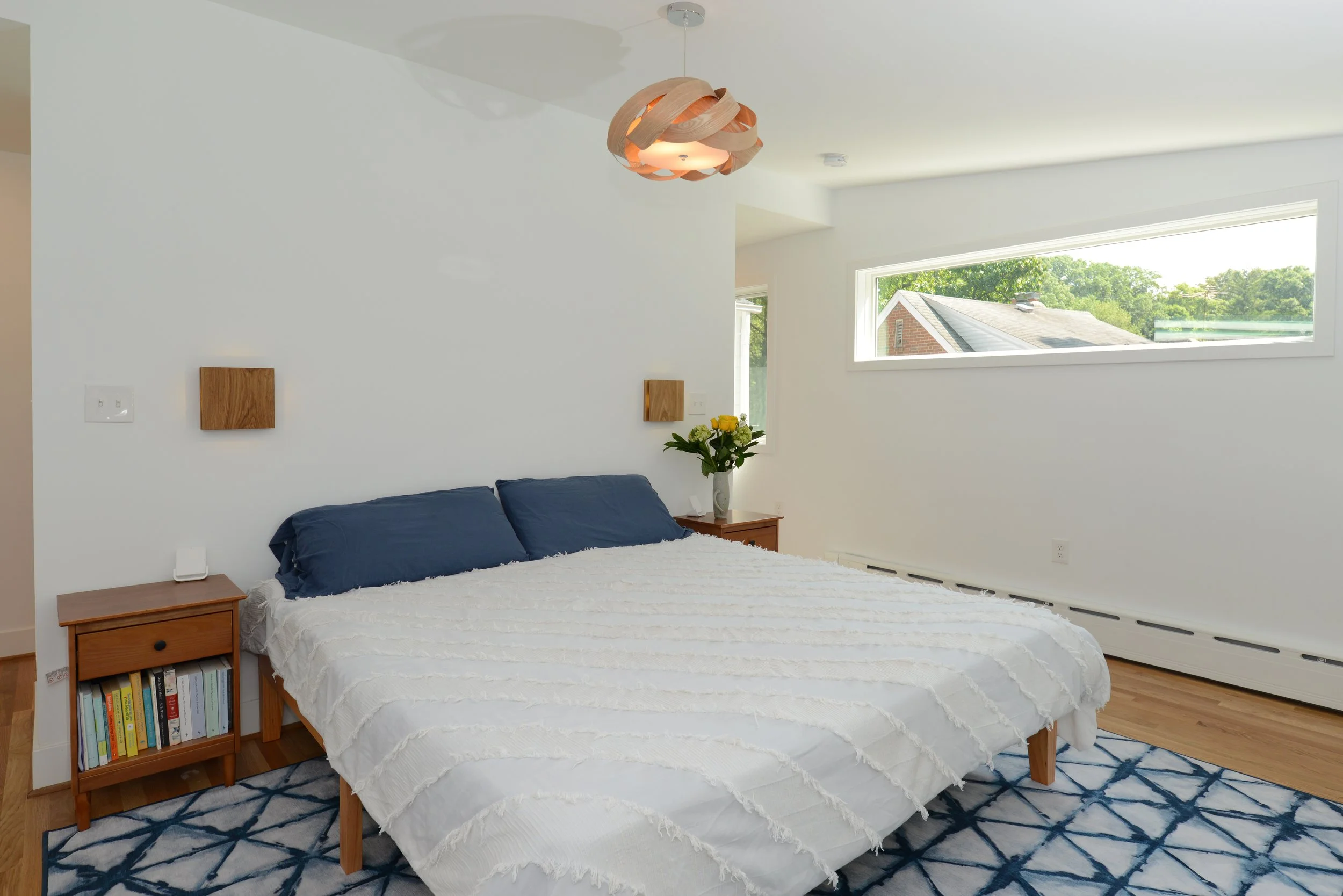 A bedroom with a bed that has dark blue pillows and a white blanket, two wooden nightstands with books and a vase of flowers, a geometric patterned rug, and a large horizontal window showing an outside view of houses and trees.