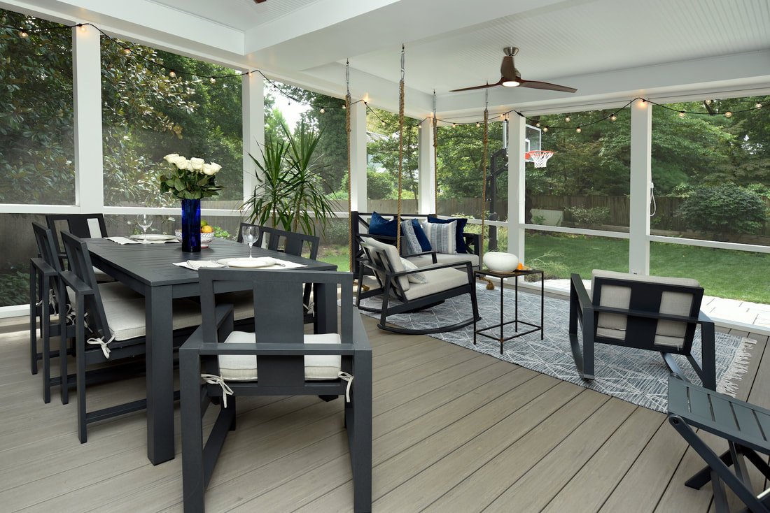 Enclosed porch with black dining table and chairs, a white cushioned rocking chair, a small side table with white decor, and string lights; surrounded by greenery and trees.