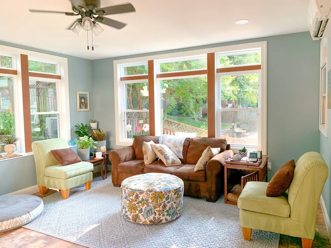 Living room with three windows, a ceiling fan, and furnished with a brown sofa, two green armchairs, an oval floral ottoman, side tables, and various potted plants.