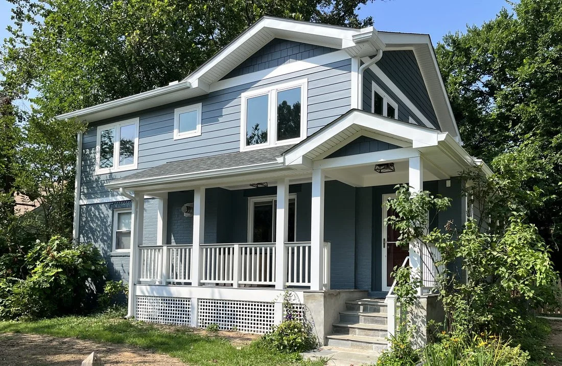 A two-story house with blue siding, white trim, and a front porch with white railings, surrounded by green trees and plants.