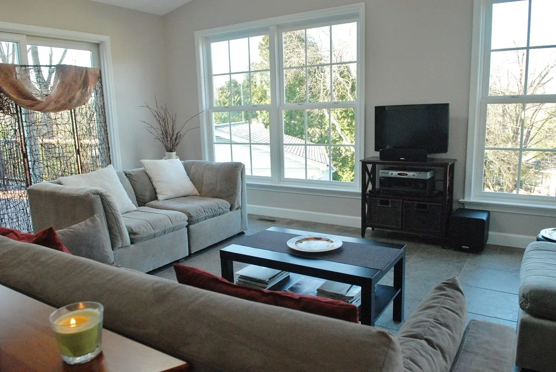 Living room with large windows, beige sofas, a black coffee table, a TV on a stand, and decorative items including a candle and a vase with branches.